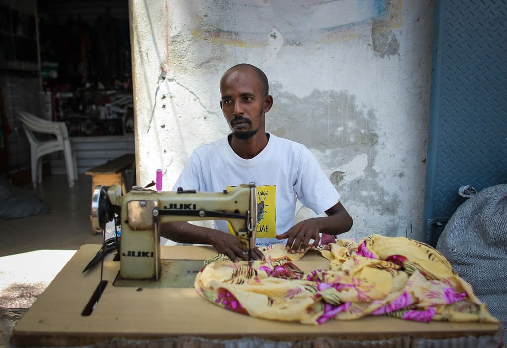 A tailor in a white T-shirt guiding yellow floral fabric through an industrial sewing machine at his stall.