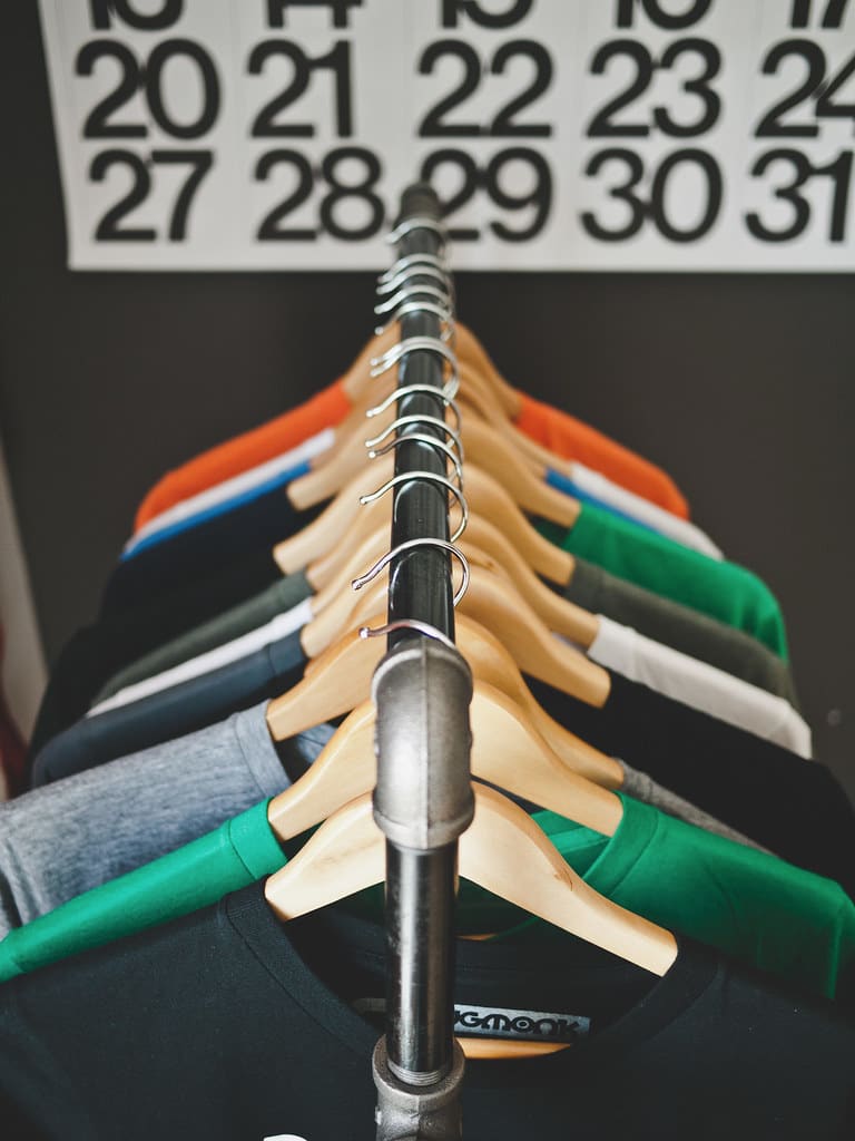 A short clothes rail of wooden hangers holding T-shirts in green, gray, white, orange and black.