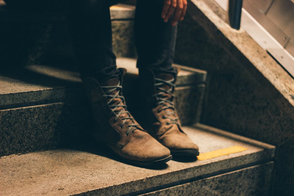 Brown leather lace-up boots worn with dark trousers on a wooden staircase, lit low from one side.