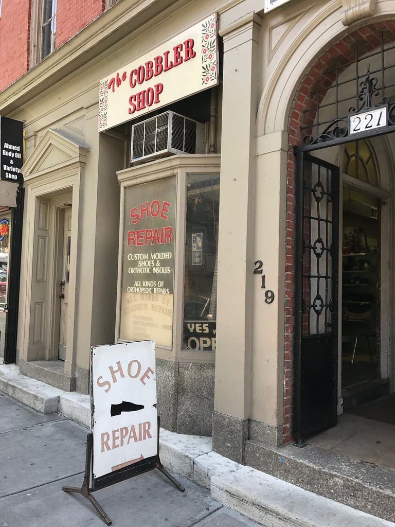 A small cobbler's storefront with "Shoe Repair" painted in the window and a sandwich board on the sidewalk.