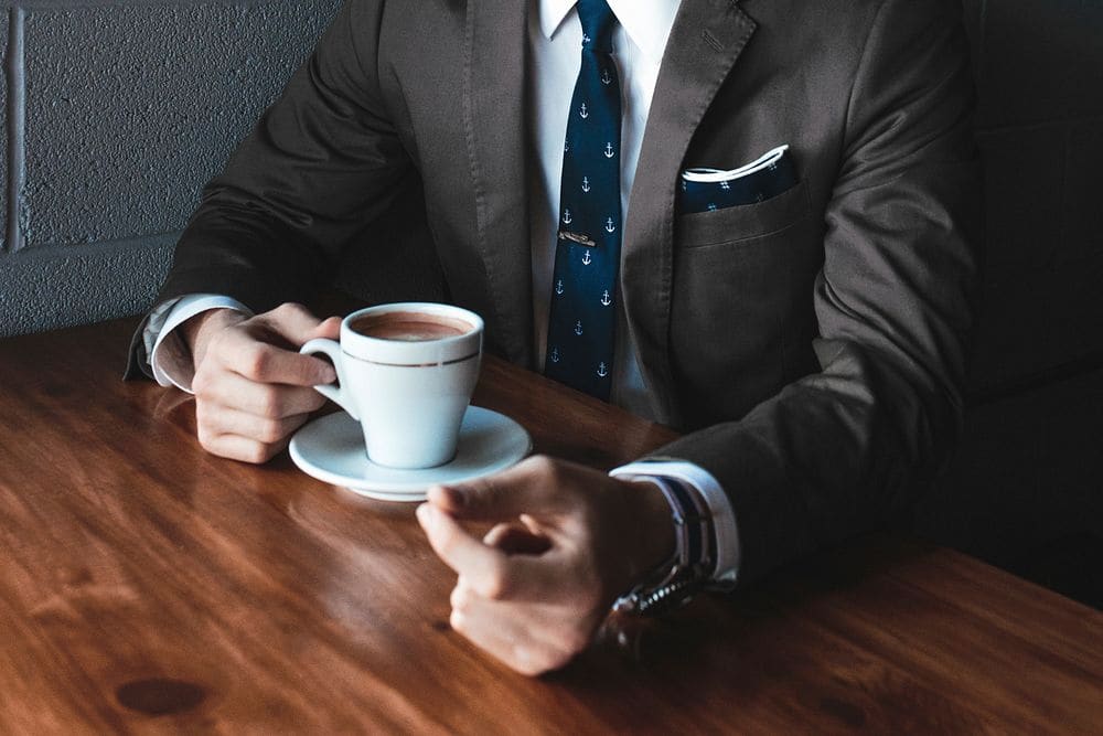 A man in a charcoal suit, white shirt and navy anchor-print tie holding a white coffee cup at a wooden table.
