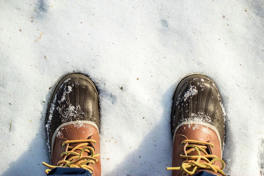 A first-person view of brown leather duck boots with rubber toes and yellow laces standing in fresh snow.