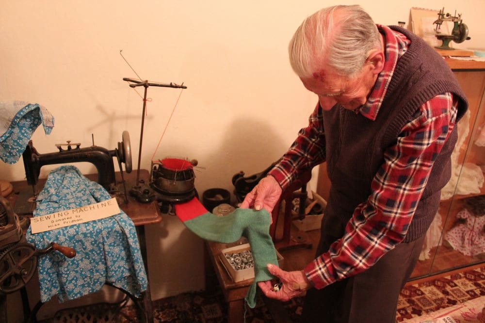 An older man in a plaid shirt and sweater vest holding a green and red sock beside an antique sock-knitting machine.