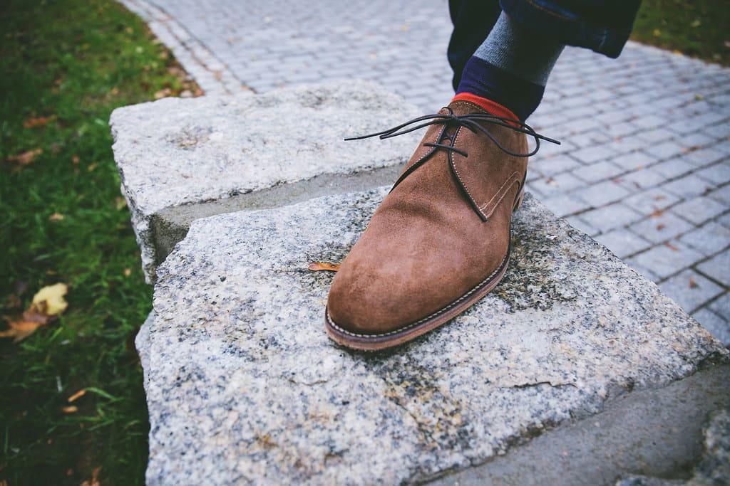 A brown suede chukka boot with dark laces resting on a block of granite beside a brick sidewalk.