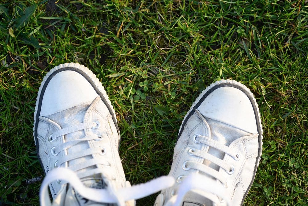 A first-person view of worn white canvas sneakers with loose laces standing in green grass.
