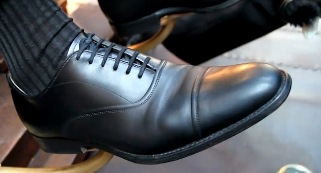 A black leather cap-toe oxford with closed lacing propped on a shoeshine stand.